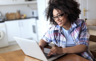 Woman with glasses sitting at her kitchen table smiling at her laptop